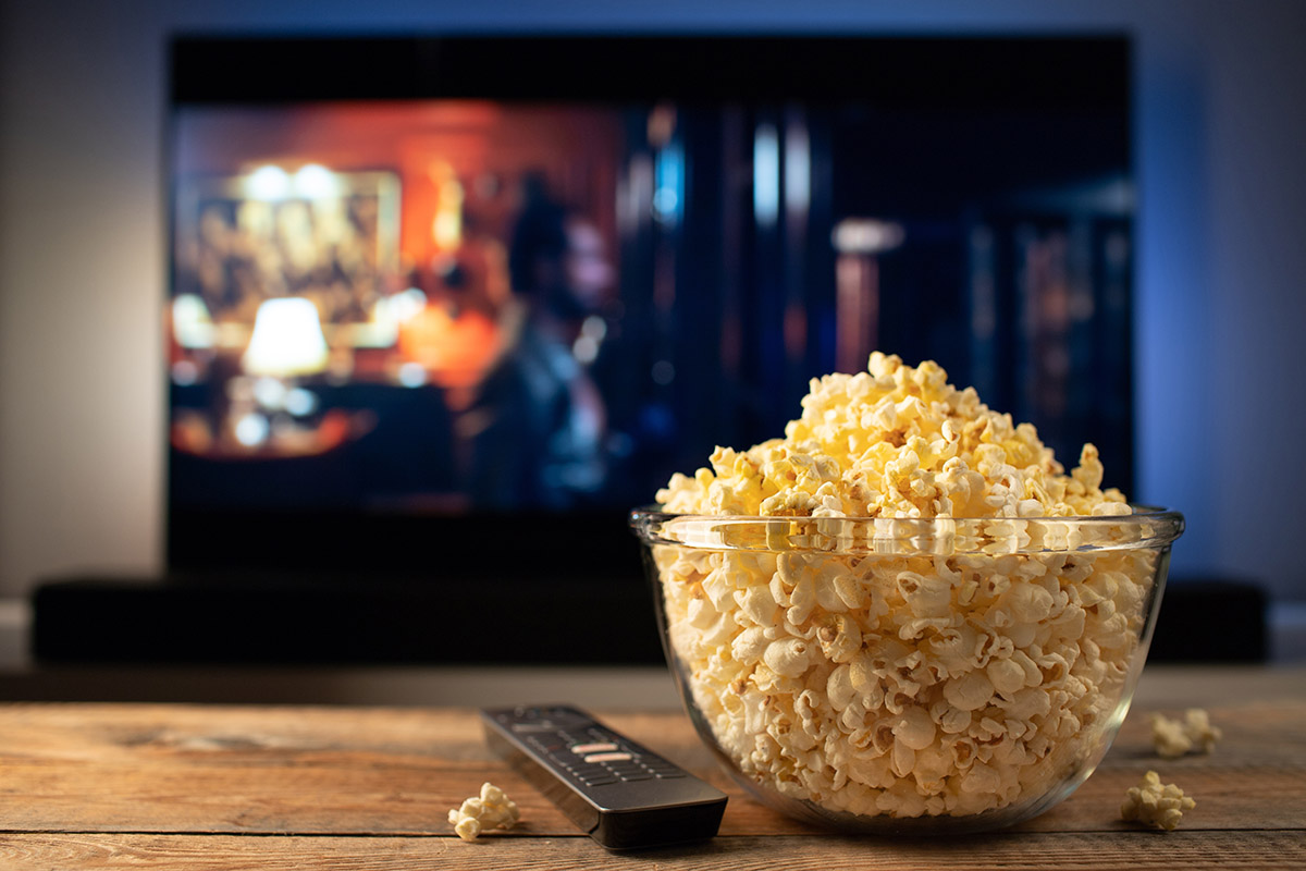 A glass bowl of popcorn and remote control in the background the TV works. Evening cozy watching a movie or TV series at home.